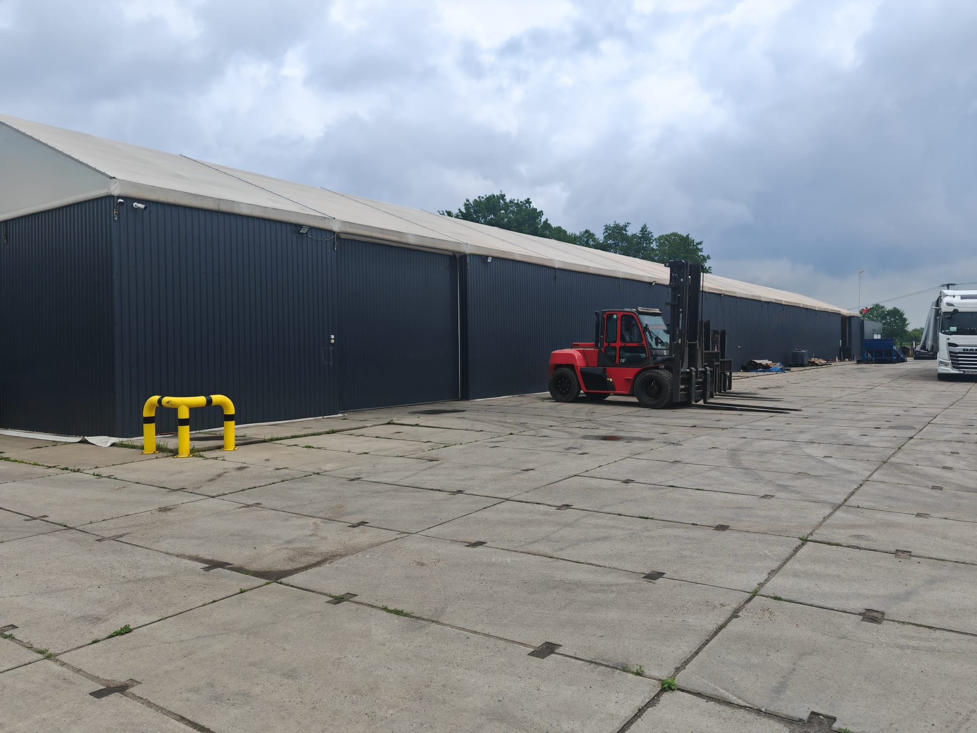 Czerwony wózek widłowy na tle długiego, czarnego magazynu z białym dachem i betonowego placu. W tle widoczny fragment białej ciężarówki.