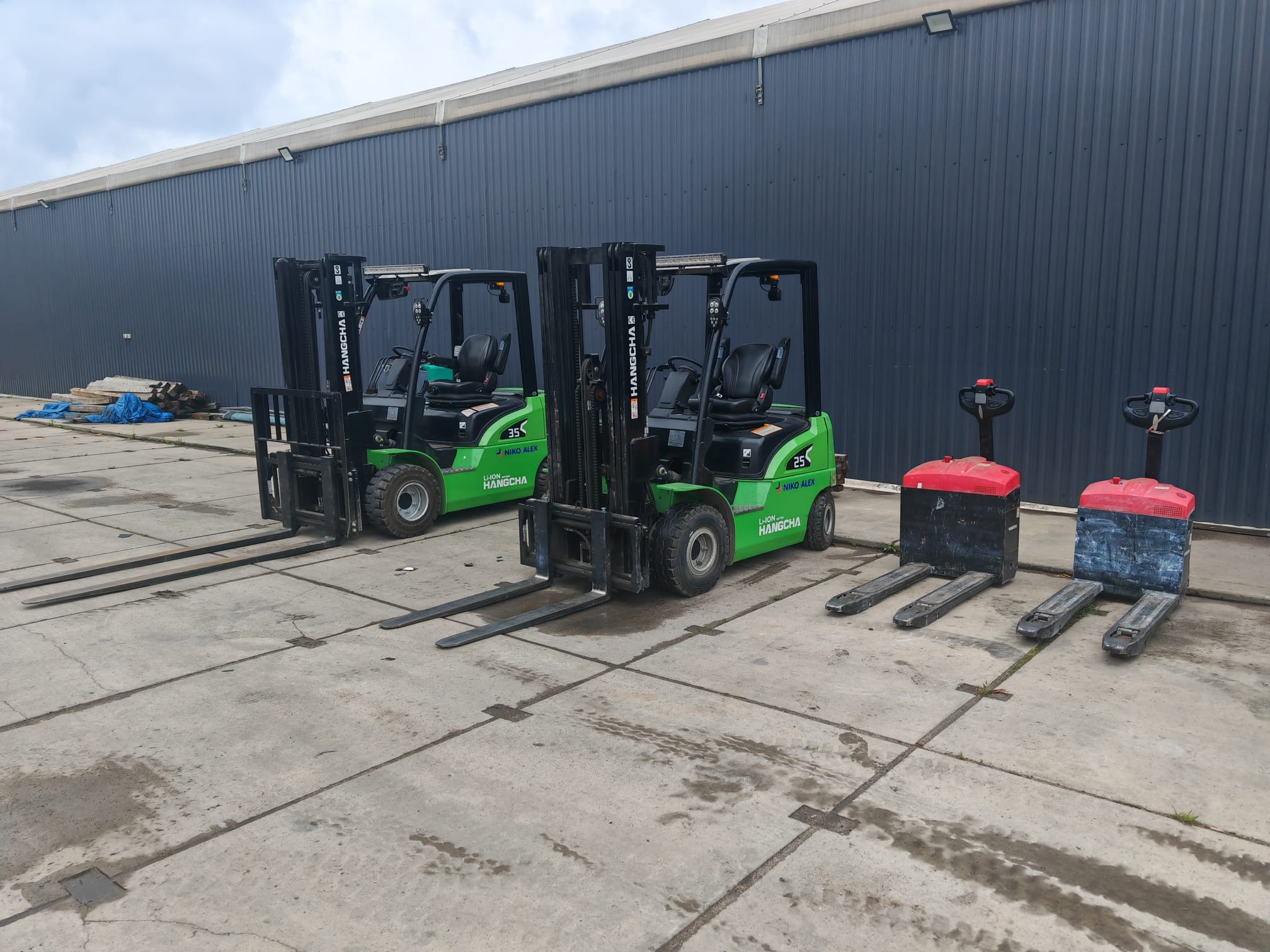 Dwa zielone wózki widłowe Hangcha i dwa wózki paletowe z czerwonymi obudowami na betonowym placu przed szarą halą magazynową, w pochmurny dzień.