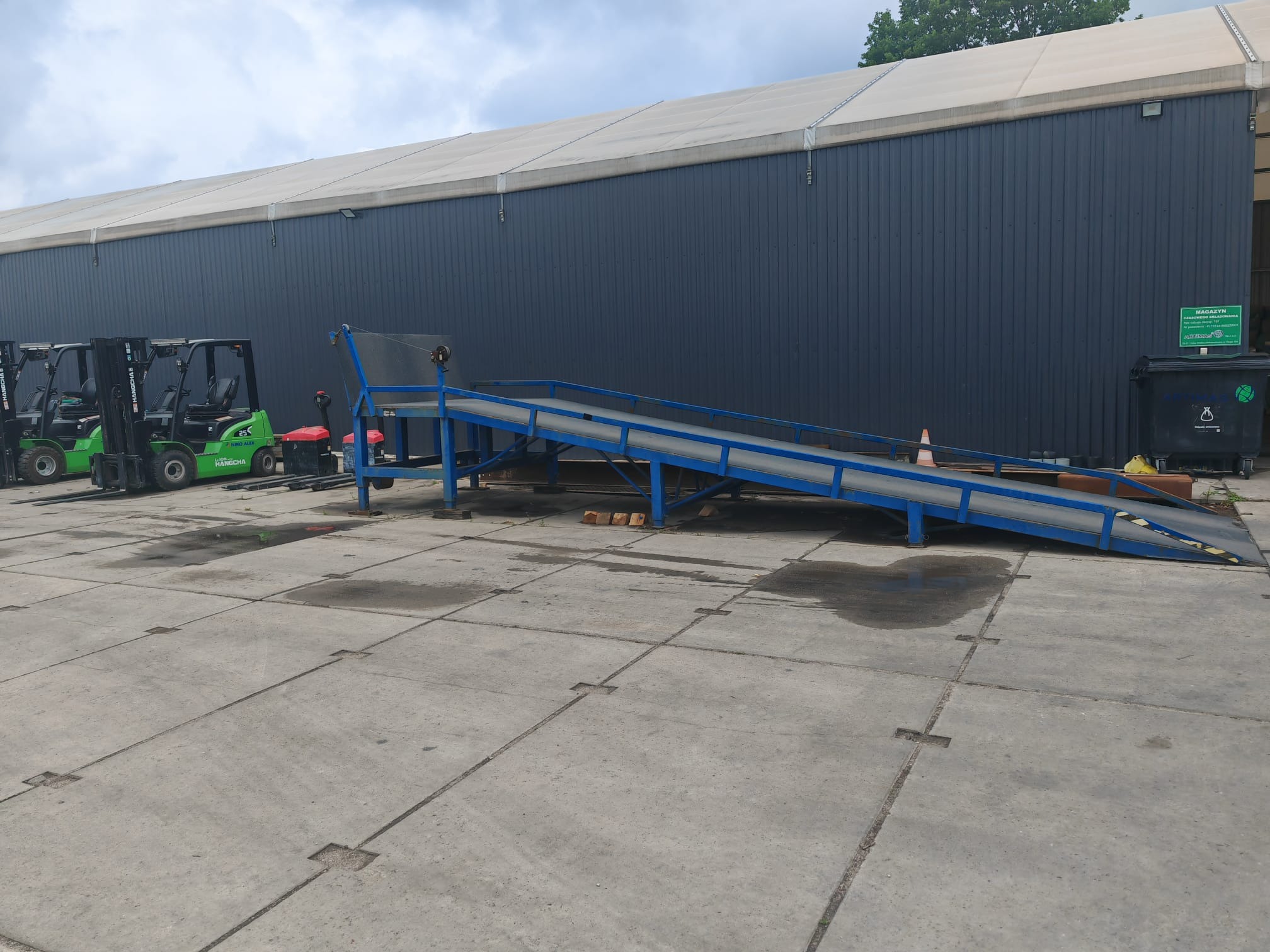 Rampa załadunkowa przed magazynem z zaparkowanymi wózkami widłowymi. Widoczny budynek magazynu w kolorze grafitowym, betonowa rampa w kolorze niebieskim.