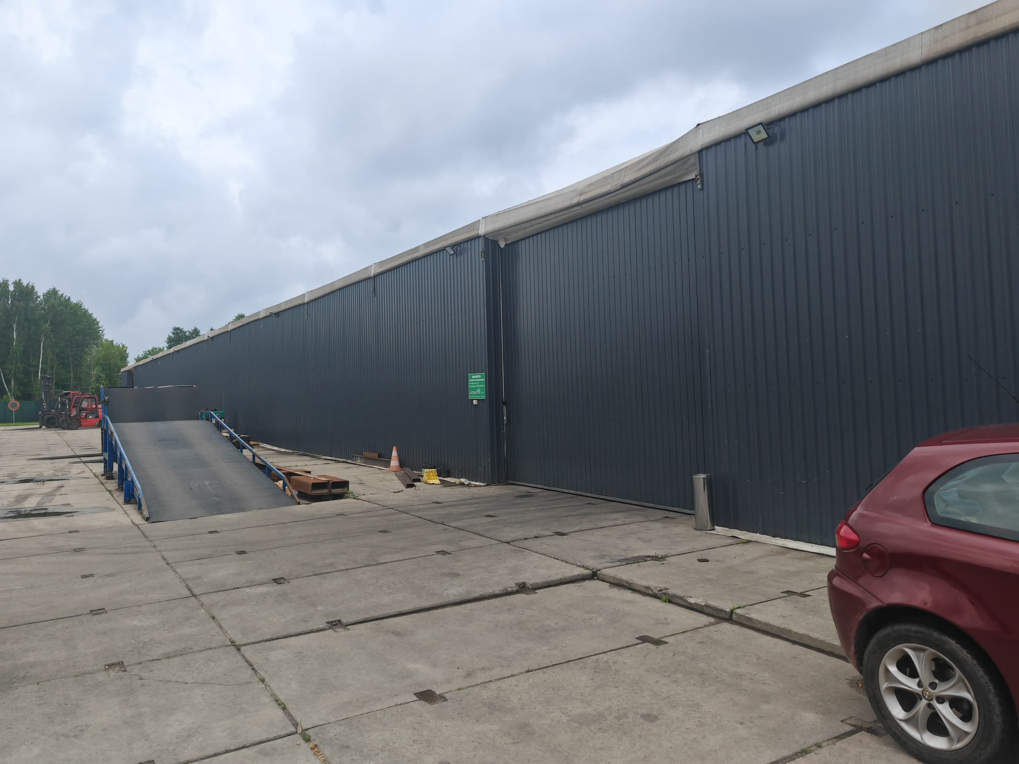 Rampa załadunkowa przy magazynie z szarej blachy falistej. Czerwony wózek widłowy i auto osobowe w tle. Betonowy plac z widocznymi spoinami.