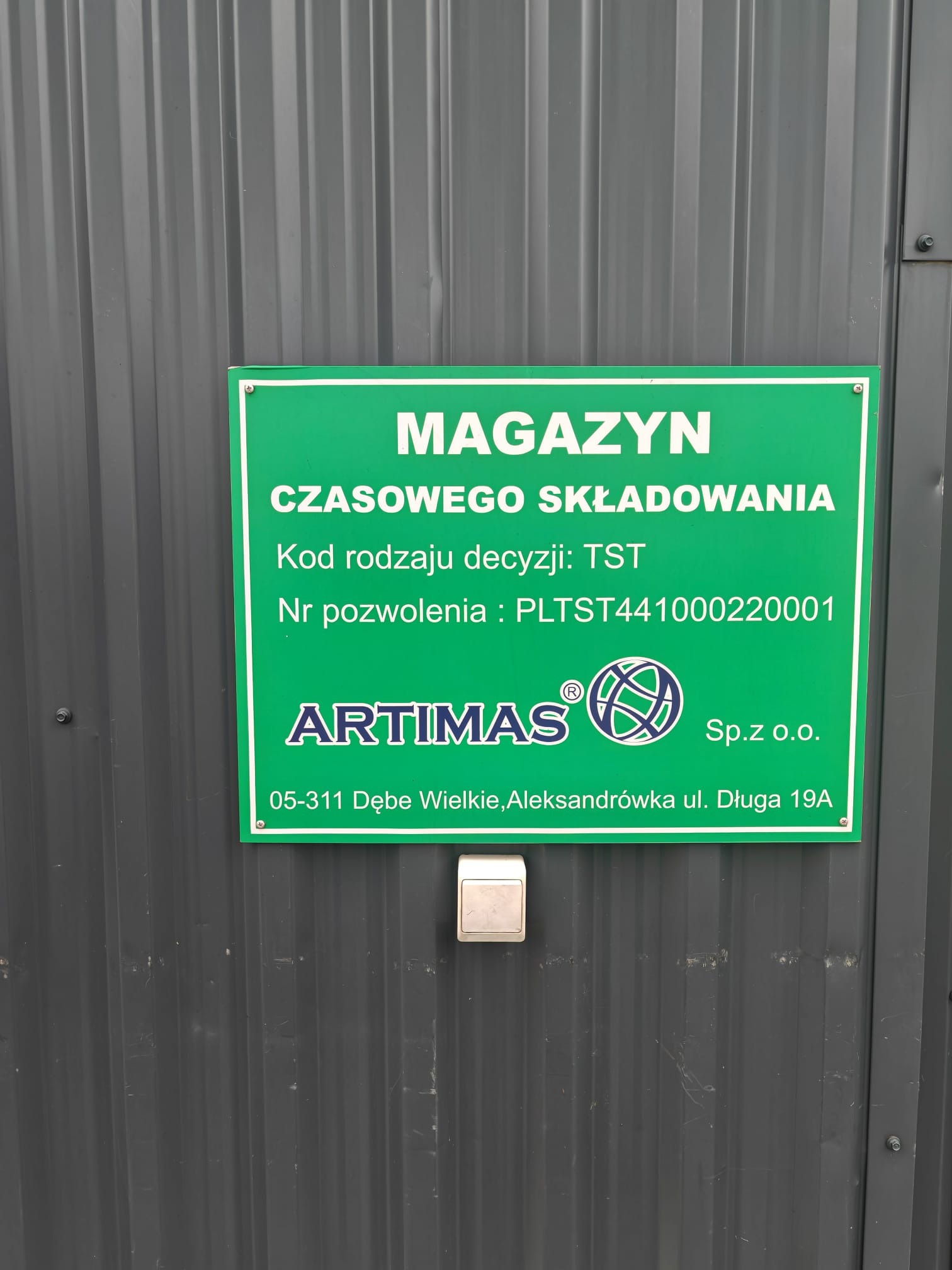 Tablica informacyjna na szarym magazynie z napisem 'Magazyn Czasowego Składowania' i danymi firmy Artimas Sp. z o.o. w Dębem Wielkim.