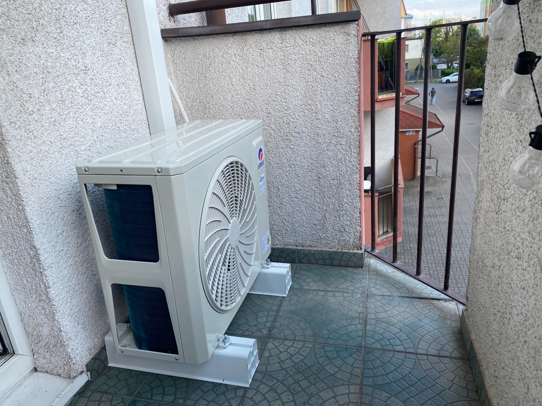 Biała jednostka zewnętrzna klimatyzacji Gree, zamontowana na balkonie z płytkami w odcieniach zieleni. Widoczne elementy mocujące i fragment zabudowy balkonu.