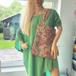 Manekin w zielonej sukience prezentuje torbę na ramię z barwnym, orientalnym wzorem. W tle okno z widokiem na ogród i rolki materiałów.