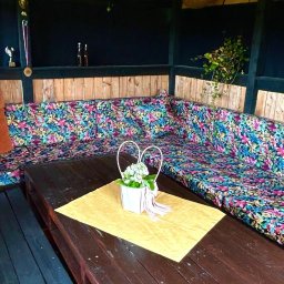 DONATA ZIELONKA USLUGI KRAWIECKIE - Wnętrze altany z narożną kanapą w kwiatowe wzory, drewnianym stołem i dekoracjami. Widoczne trofea na półce i kosze pod sufitem.