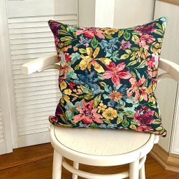 DONATA ZIELONKA USLUGI KRAWIECKIE - Kwadratowa poduszka dekoracyjna z ciemnym tłem i wzorem w kolorowe kwiaty leży na białym krześle w jasnym wnętrzu. Widoczna podłoga z paneli i szafka.