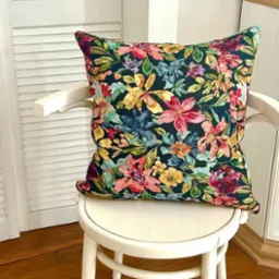 Kwadratowa poduszka dekoracyjna z ciemnym tłem i wzorem w kolorowe kwiaty leży na białym krześle w jasnym wnętrzu. Widoczna podłoga z paneli i szafka.