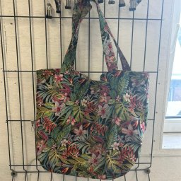 DONATA ZIELONKA USLUGI KRAWIECKIE - Tkaninowa torba na zakupy z motywem tropikalnych liści i kwiatów, zawieszona na metalowym wieszaku z klipsami, widok z przodu.