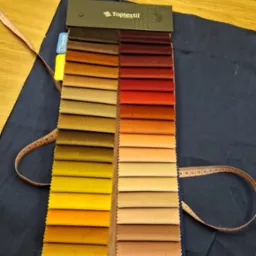 Próbnik tkanin Toptextil z paletą kolorów na ciemnoniebieskim materiale, obok leży miarka krawiecka i inne akcesoria krawieckie na drewnianym stole.