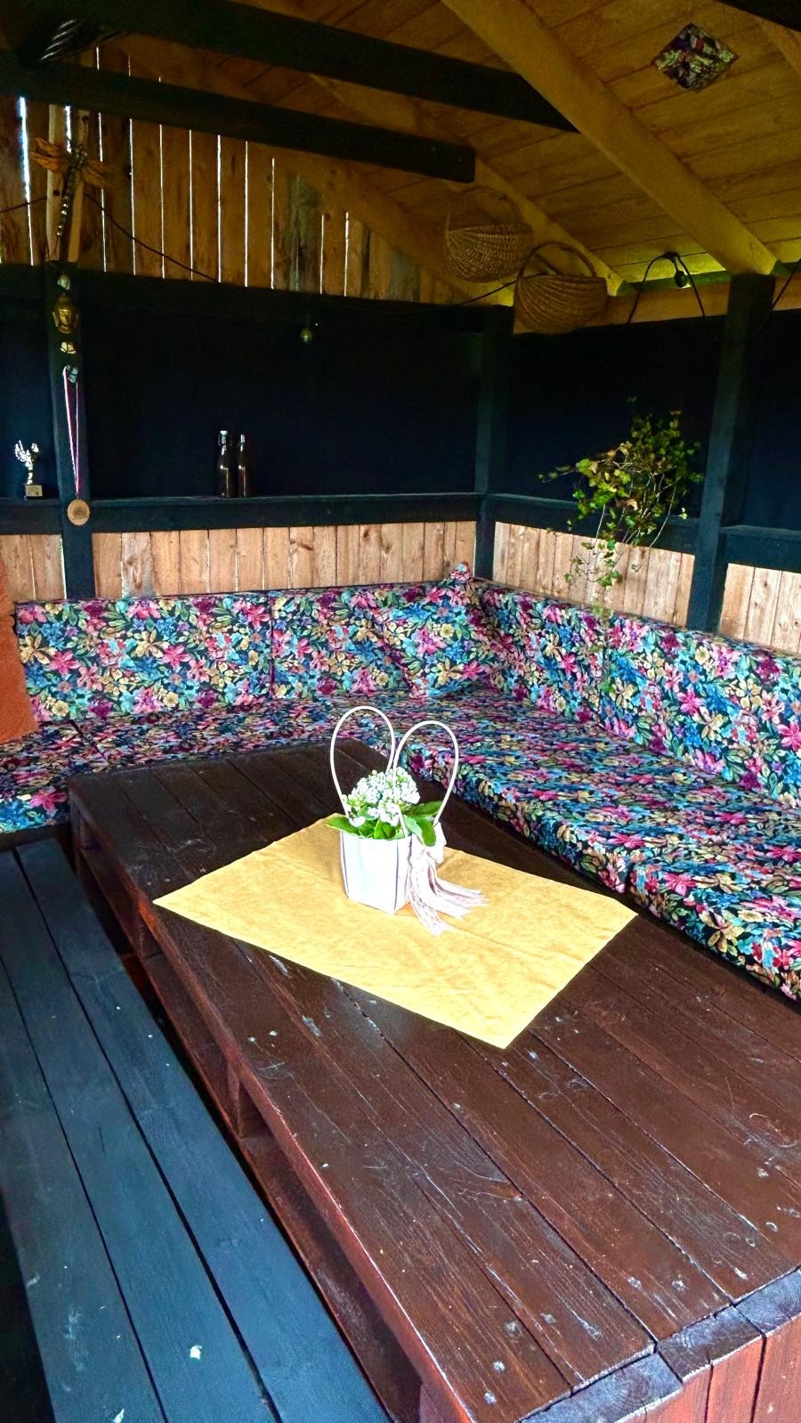 Wnętrze altany z narożną kanapą w kwiatowe wzory, drewnianym stołem i dekoracjami. Widoczne trofea na półce i kosze pod sufitem.