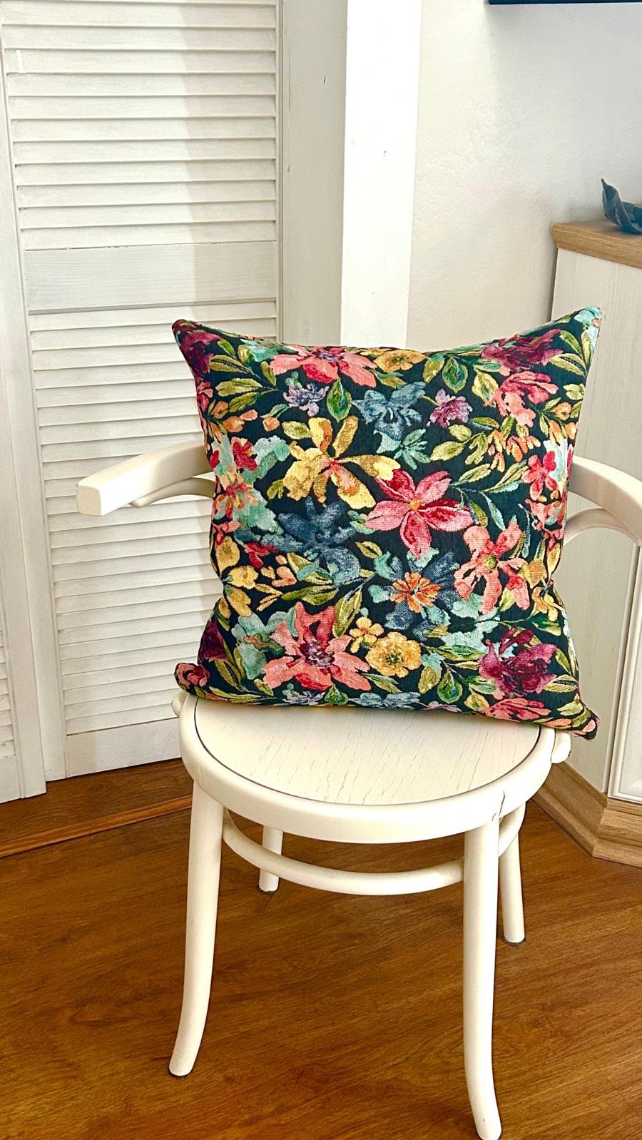 Kwadratowa poduszka dekoracyjna z ciemnym tłem i wzorem w kolorowe kwiaty leży na białym krześle w jasnym wnętrzu. Widoczna podłoga z paneli i szafka.