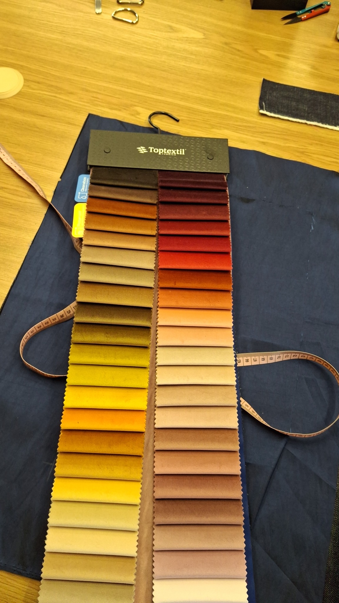 Próbnik tkanin Toptextil z paletą kolorów na ciemnoniebieskim materiale, obok leży miarka krawiecka i inne akcesoria krawieckie na drewnianym stole.