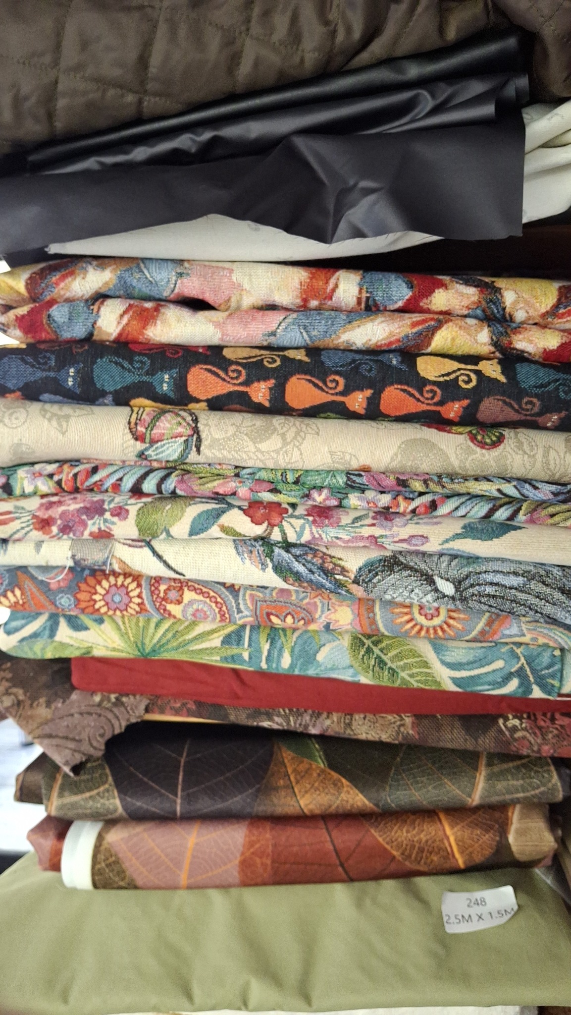 Stos różnorodnych materiałów tekstylnych, w tym tkaniny wzorzyste, skóra ekologiczna i pikowany materiał, ułożonych jeden na drugim. Widoczna metka z wymiarami.