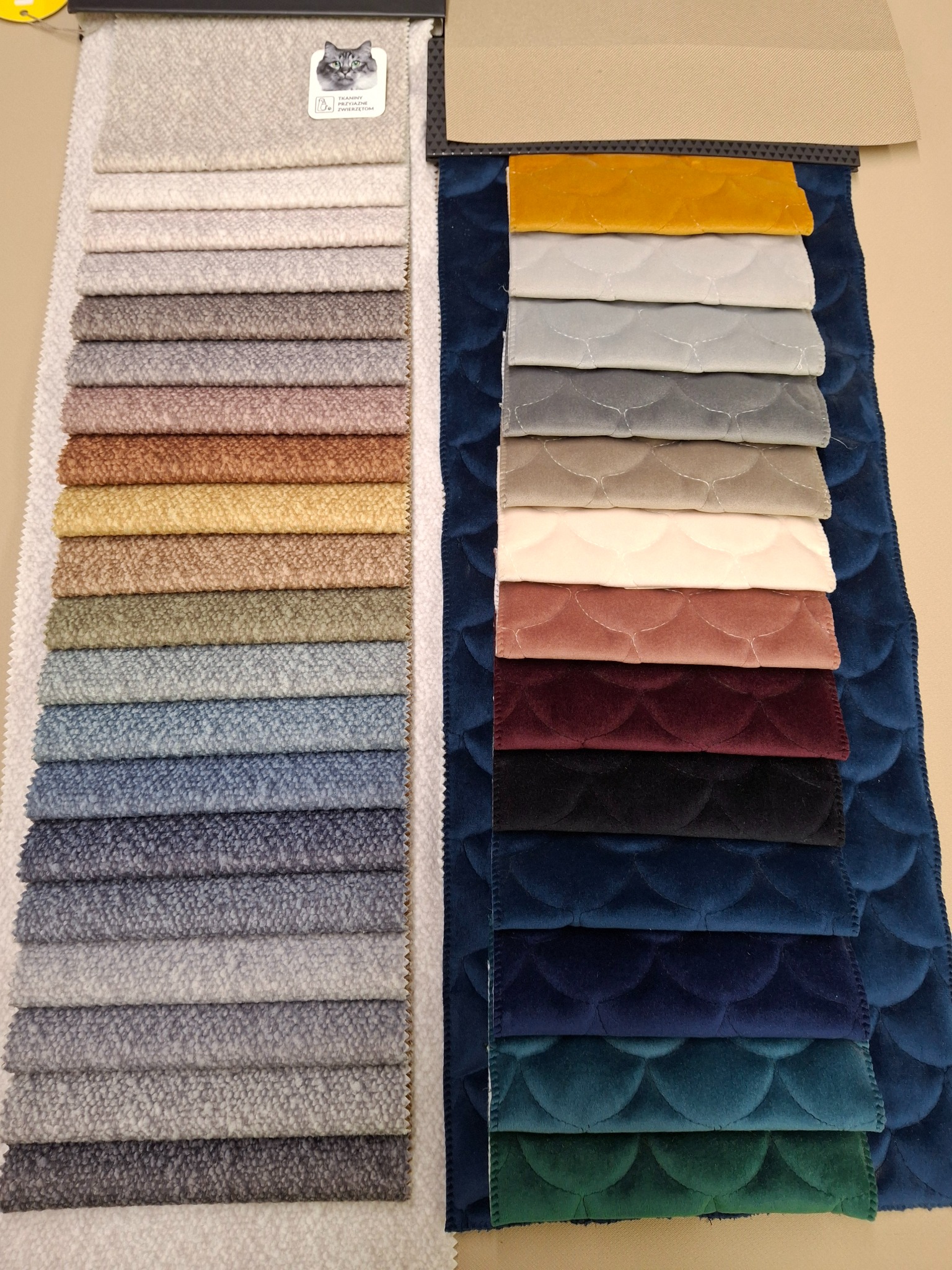 Próbki tkanin tapicerskich w różnych kolorach i fakturach, ułożone obok siebie. Widoczne materiały gładkie i z wyraźną strukturą, paleta barw od jasnych szarości po głębokie odcienie granatu.