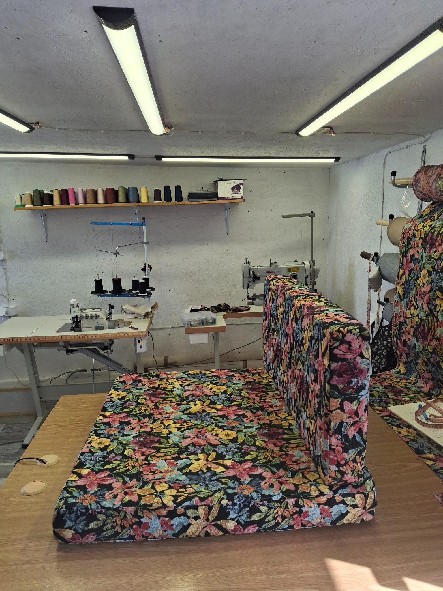 Tapicerowany zagłówek łóżka w kwiatowy wzór, w warsztacie. Widoczne maszyny do szycia, nici i materiały. Oświetlenie jarzeniowe, drewniany stół roboczy.
