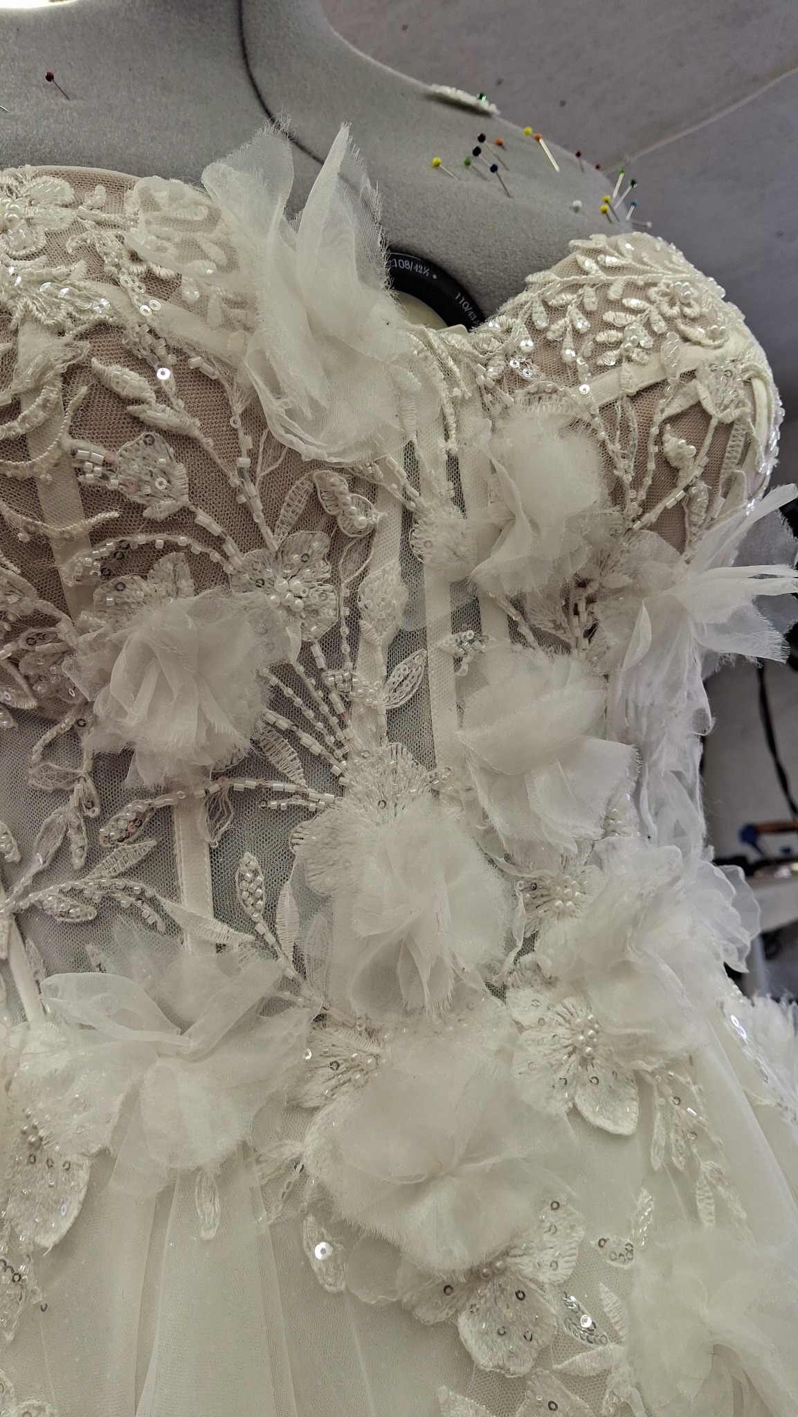 Biała suknia ślubna z koronkowymi aplikacjami i kwiatowymi zdobieniami na manekinie krawieckim, widoczne szpilki, detale wykończenia.
