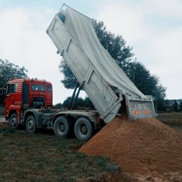 SDB Zenon Młynek - Czerwona ciężarówka z otwartą naczepą wysypuje brązowawy materiał na ziemię. W tle drzewa i budynki.