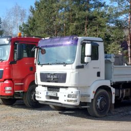 SDB Zenon Młynek - Trzy ciężarówki MAN w kolorach turkusowym, czerwonym i białym, zaparkowane na żwirowym terenie z drzewami w tle. Widoczne rury betonowe i opony.