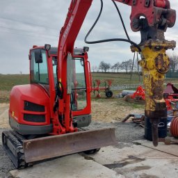 SDB Zenon Młynek - Czerwona minikoparka Volvo z hydraulicznym młotem udarowym na betonowej płycie. W tle pole i sprzęt rolniczy pod pochmurnym niebem.