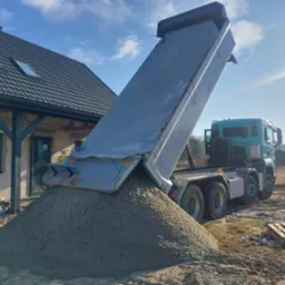 Ciężarówka wyładowuje piasek na działce budowlanej obok domu w trakcie budowy, w słoneczny dzień. Widoczny fragment dachu i okna budynku.