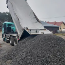 Ciężarówka z naczepą wyładowuje dużą górę ciemnego żwiru na podwórku. W tle domy i drzewa. Niebo pochmurne.