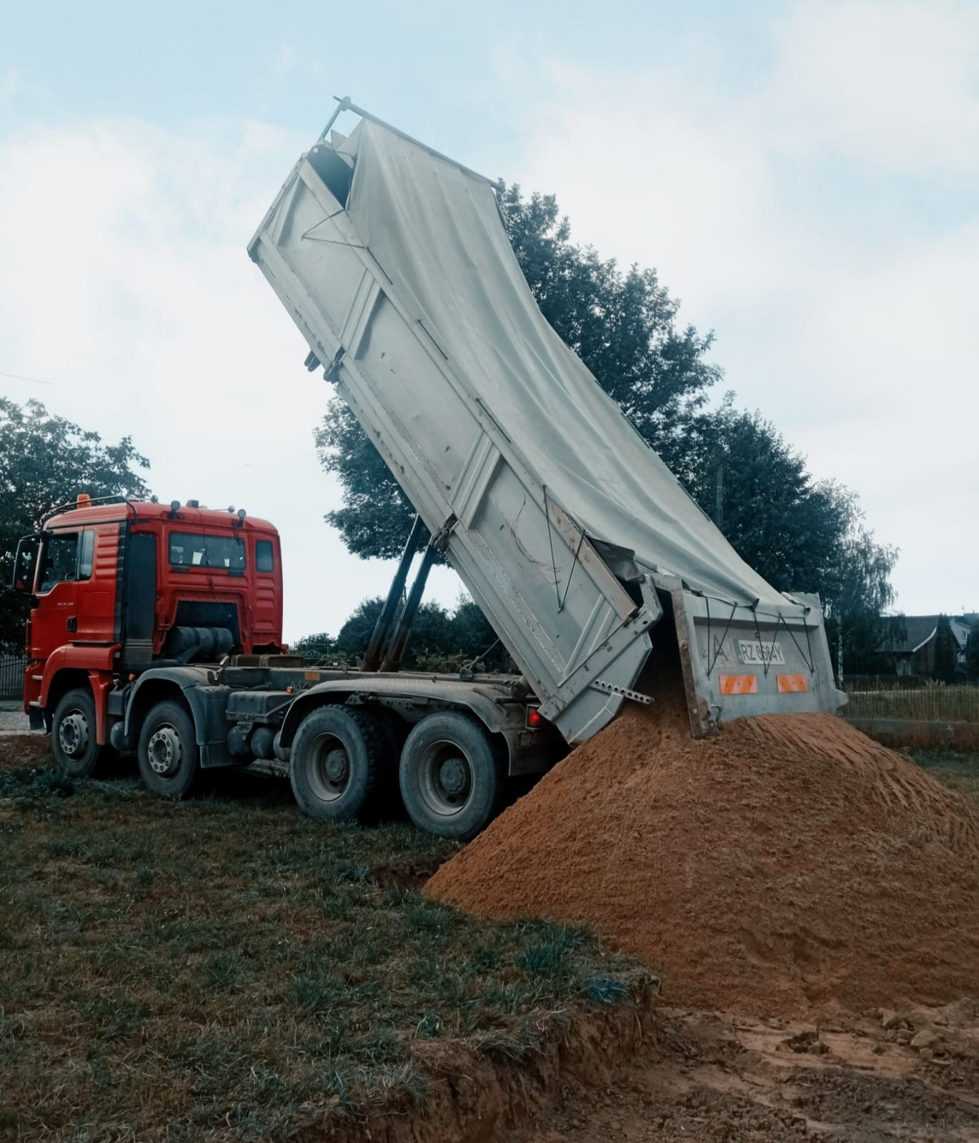 Czerwona ciężarówka z otwartą naczepą wysypuje brązowawy materiał na ziemię. W tle drzewa i budynki.