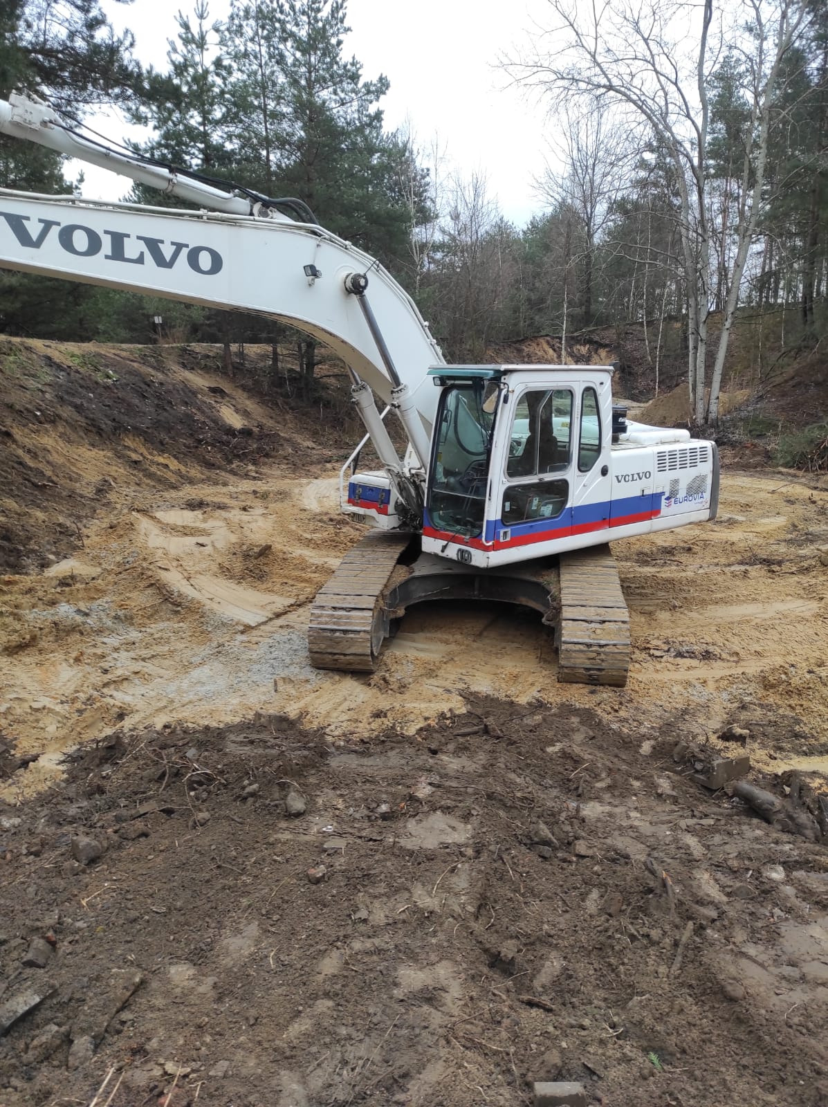 Biała koparka Volvo na placu budowy z wykopaną ziemią, widoczne ślady gąsienic i drzewa w tle. Prace ziemne w toku, dzień pochmurny.