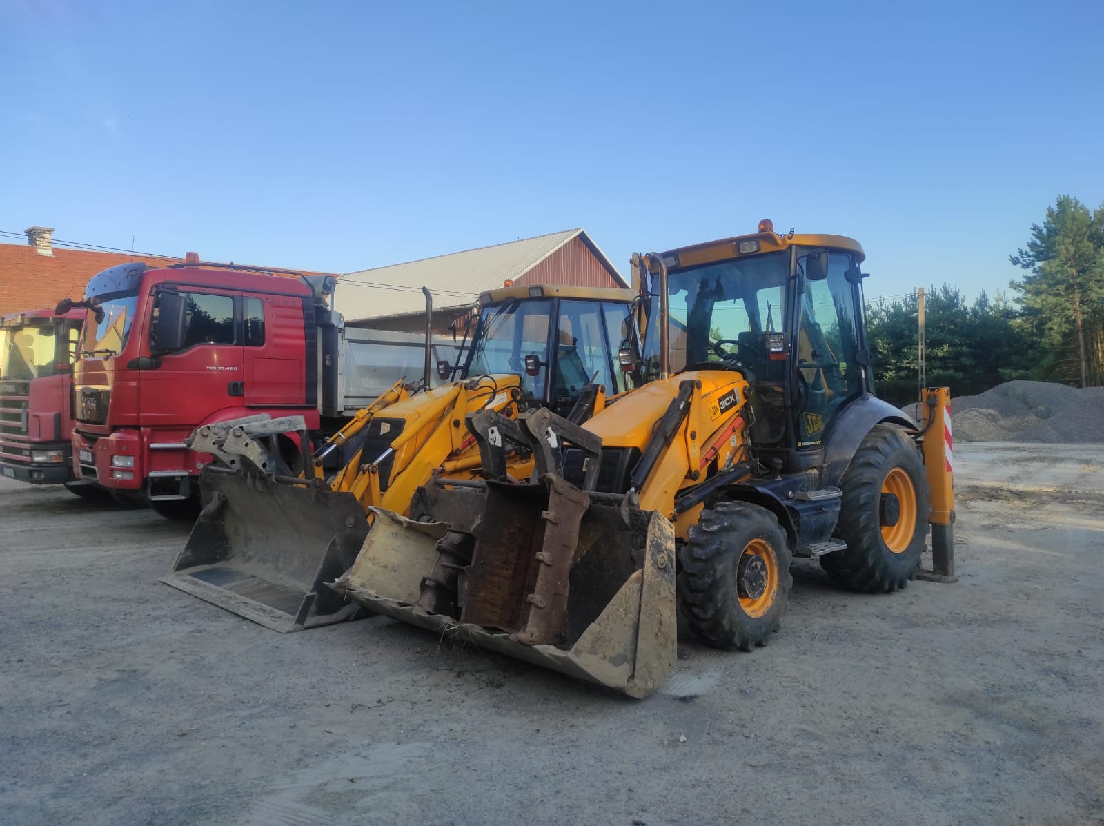 Ciężarówki i koparko-ładowarki JCB na placu budowy, z widocznym żwirem w tle, sugerujące prace ziemne i transport materiałów budowlanych.