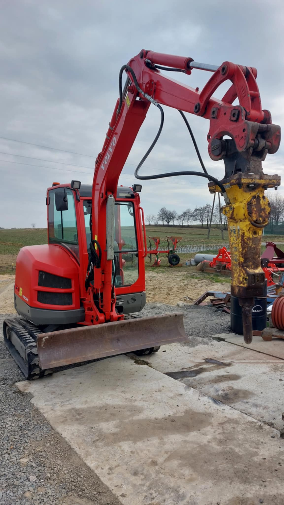 Czerwona minikoparka Volvo z hydraulicznym młotem udarowym na betonowej płycie. W tle pole i sprzęt rolniczy pod pochmurnym niebem.