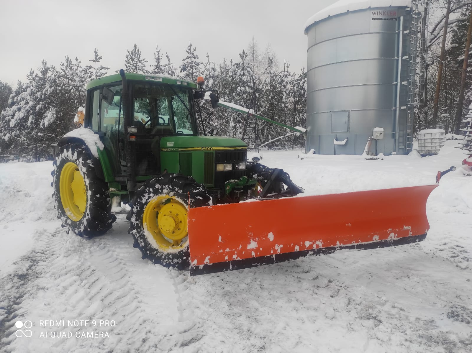 Zielony traktor John Deere 6300 z pomarańczowym pługiem odśnieżnym na tle ośnieżonego krajobrazu i metalowego silosu. Widoczne ślady opon w śniegu.