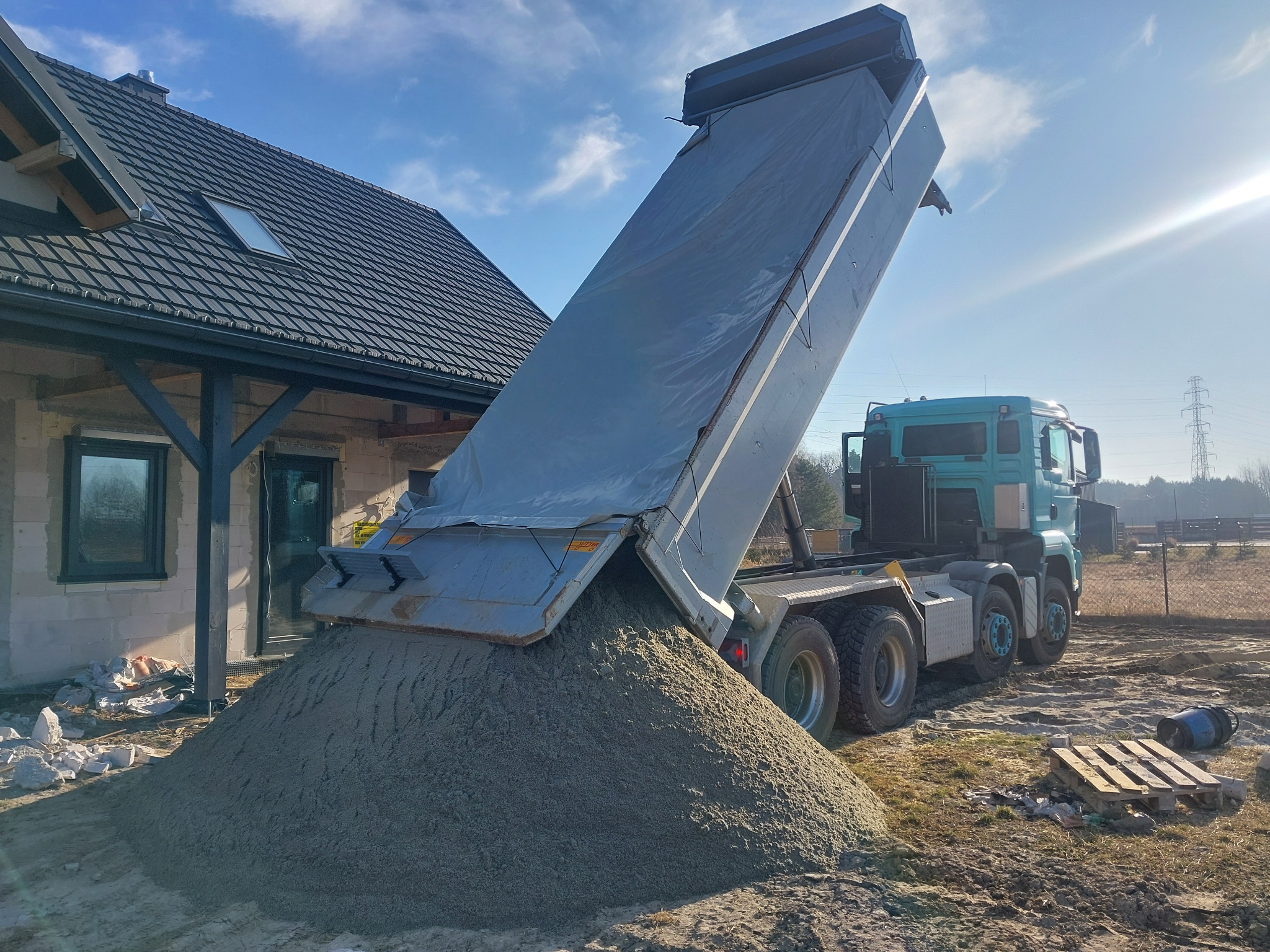 Ciężarówka wyładowuje piasek na działce budowlanej obok domu w trakcie budowy, w słoneczny dzień. Widoczny fragment dachu i okna budynku.