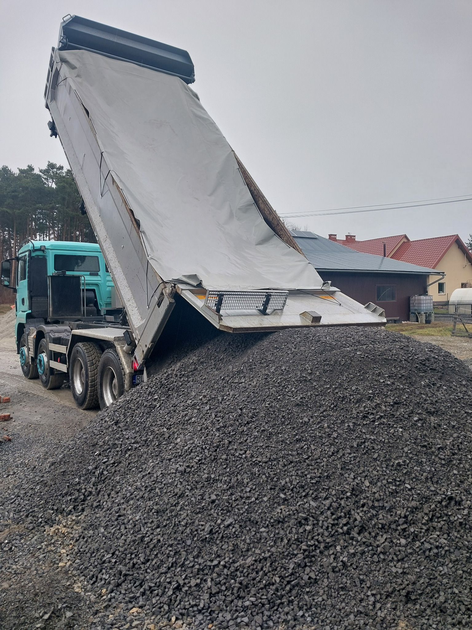Ciężarówka z naczepą wyładowuje dużą górę ciemnego żwiru na podwórku. W tle domy i drzewa. Niebo pochmurne.