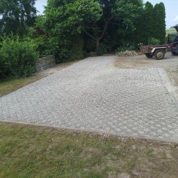 Lucjan Styrcz - Ułożona szara kostka ażurowa na podjeździe, obrzeża z kostki betonowej. W tle zieleń ogrodu i częściowo widoczny samochód z przyczepką.