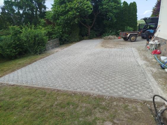 Ułożona szara kostka ażurowa na podjeździe, obrzeża z kostki betonowej. W tle zieleń ogrodu i częściowo widoczny samochód z przyczepką.