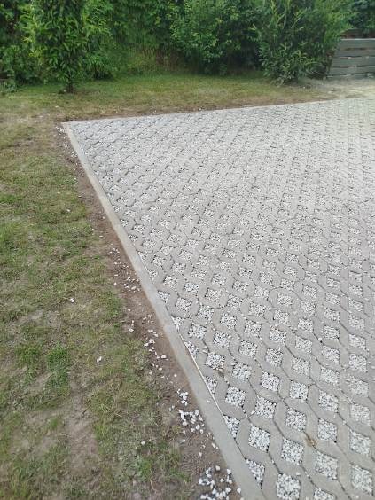 Szara, ażurowa kostka brukowa wypełniona drobnym kamieniem, oddzielona obrzeżem od trawnika. Widok z góry, lekki kąt.