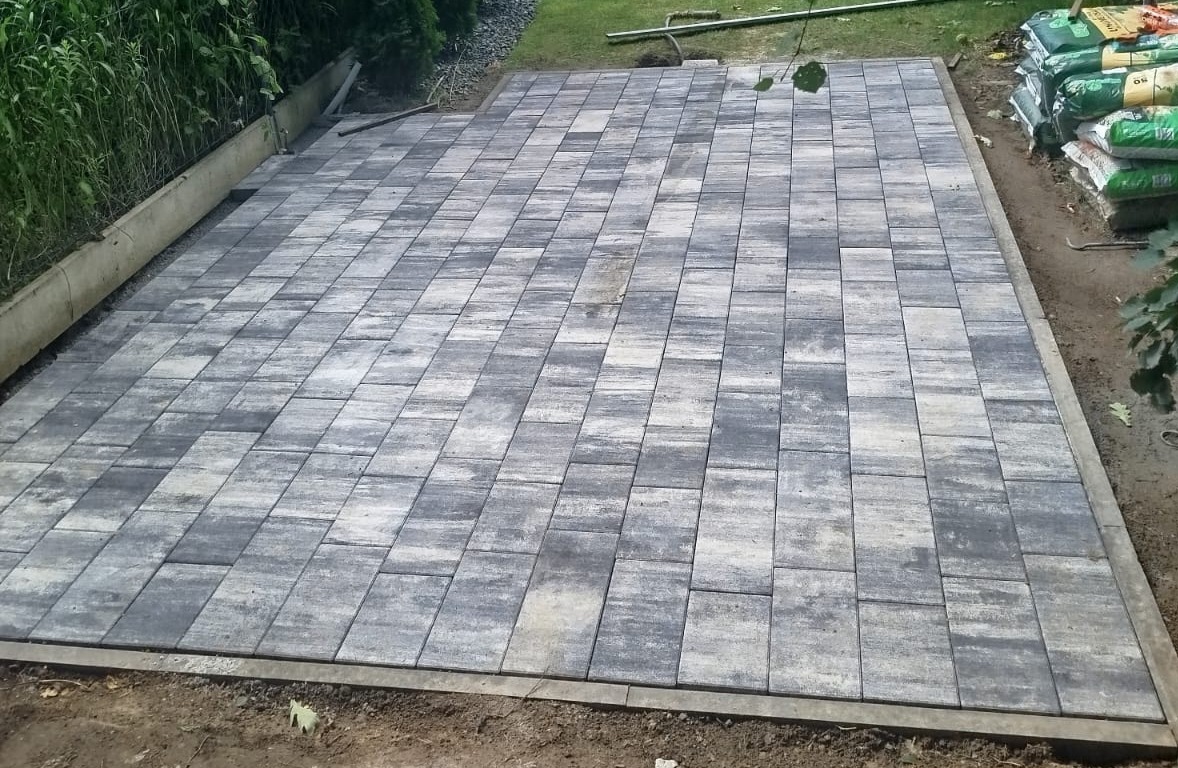 Ułożona szara kostka brukowa na posesji, widoczne worki z materiałami budowlanymi. Krawężnik oddziela kostkę od trawnika. Widok z góry na gotową nawierzchnię.
