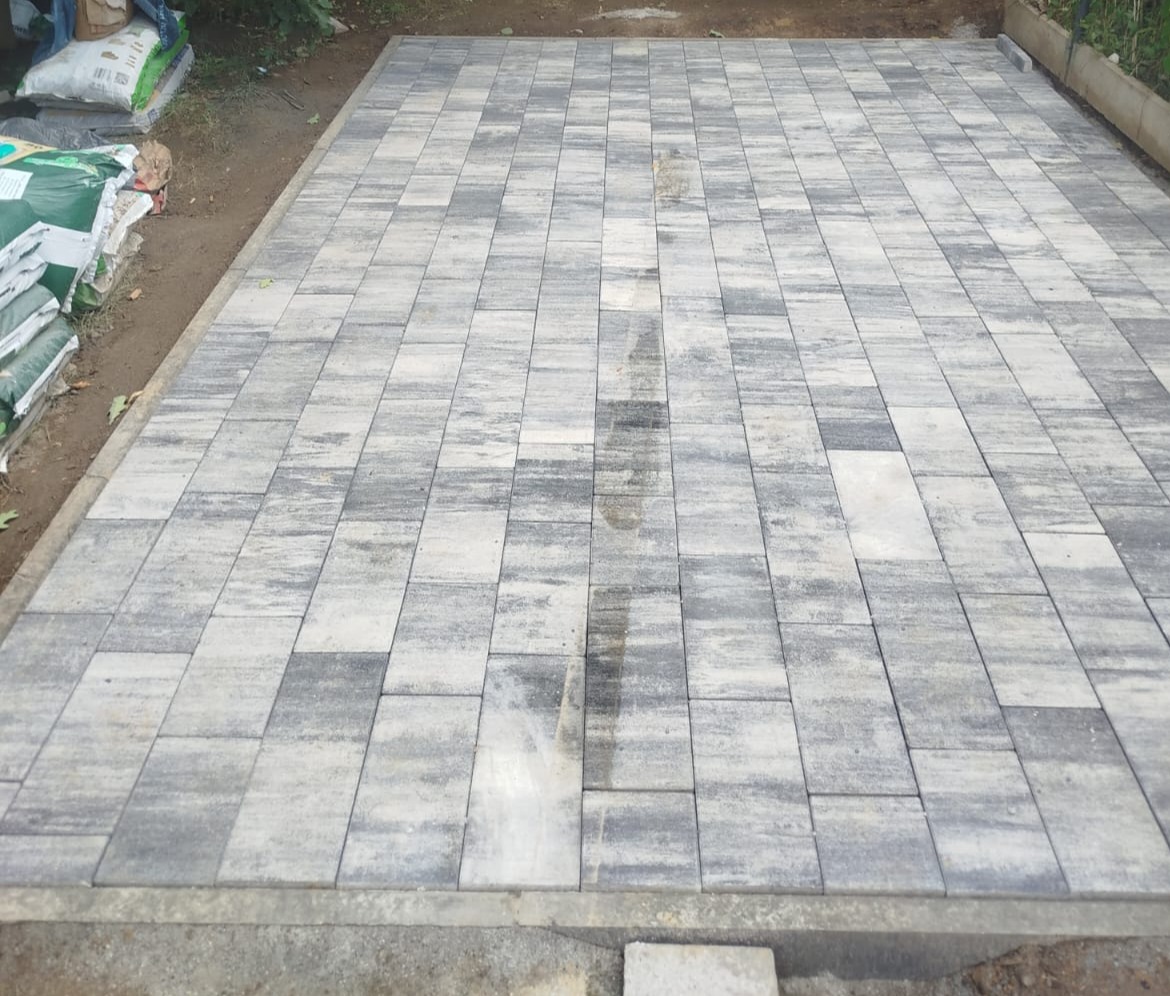 Ułożona, prostokątna kostka brukowa w odcieniach szarości i bieli, widoczna z góry. W tle worki z materiałami budowlanymi. Świeżo położona, lekko wilgotna powierzchnia.