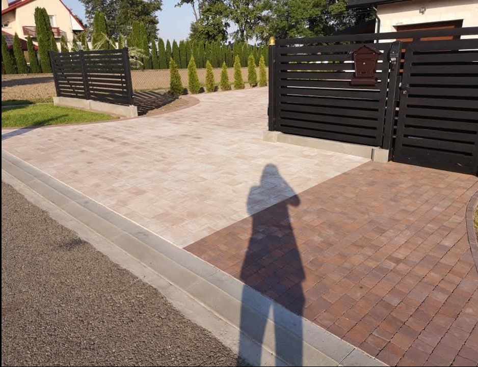 Ułożona kostka brukowa w dwóch odcieniach prowadząca do furtki z brązową skrzynką na listy. W tle rząd tuj i fragment domu.