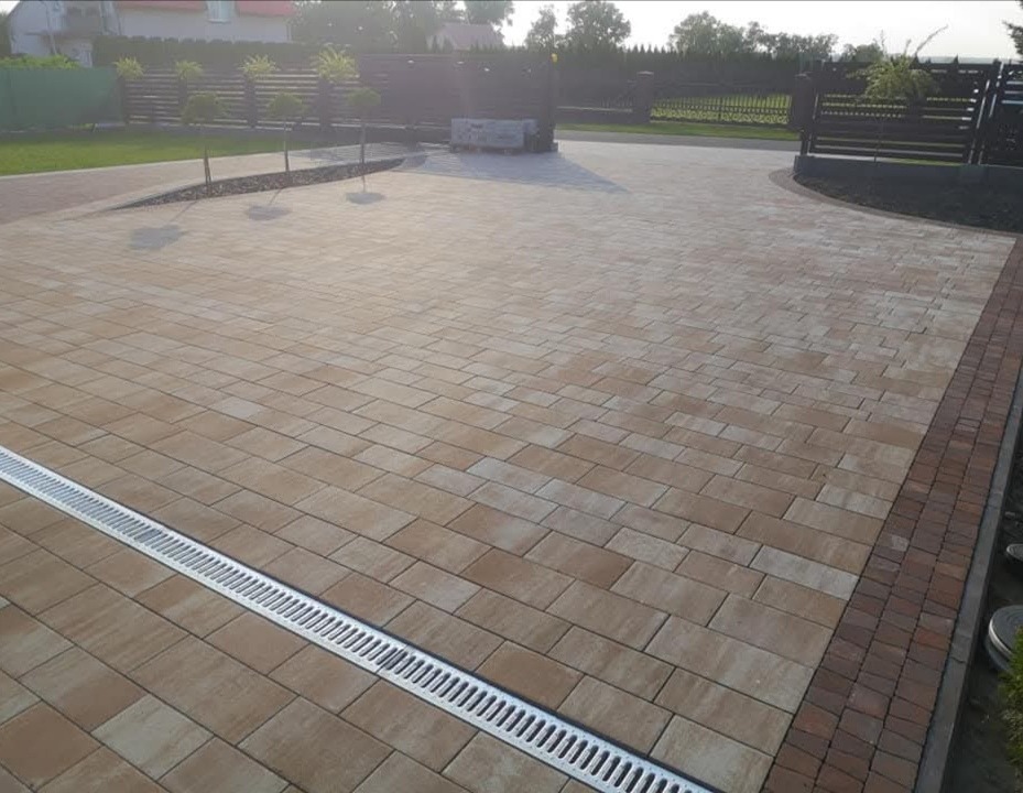Beżowa kostka brukowa z odpływem liniowym na posesji. Ciemnobrązowe obrzeża i ogrodzenie w tle. Ułożona kostka prowadzi do bramy wjazdowej.