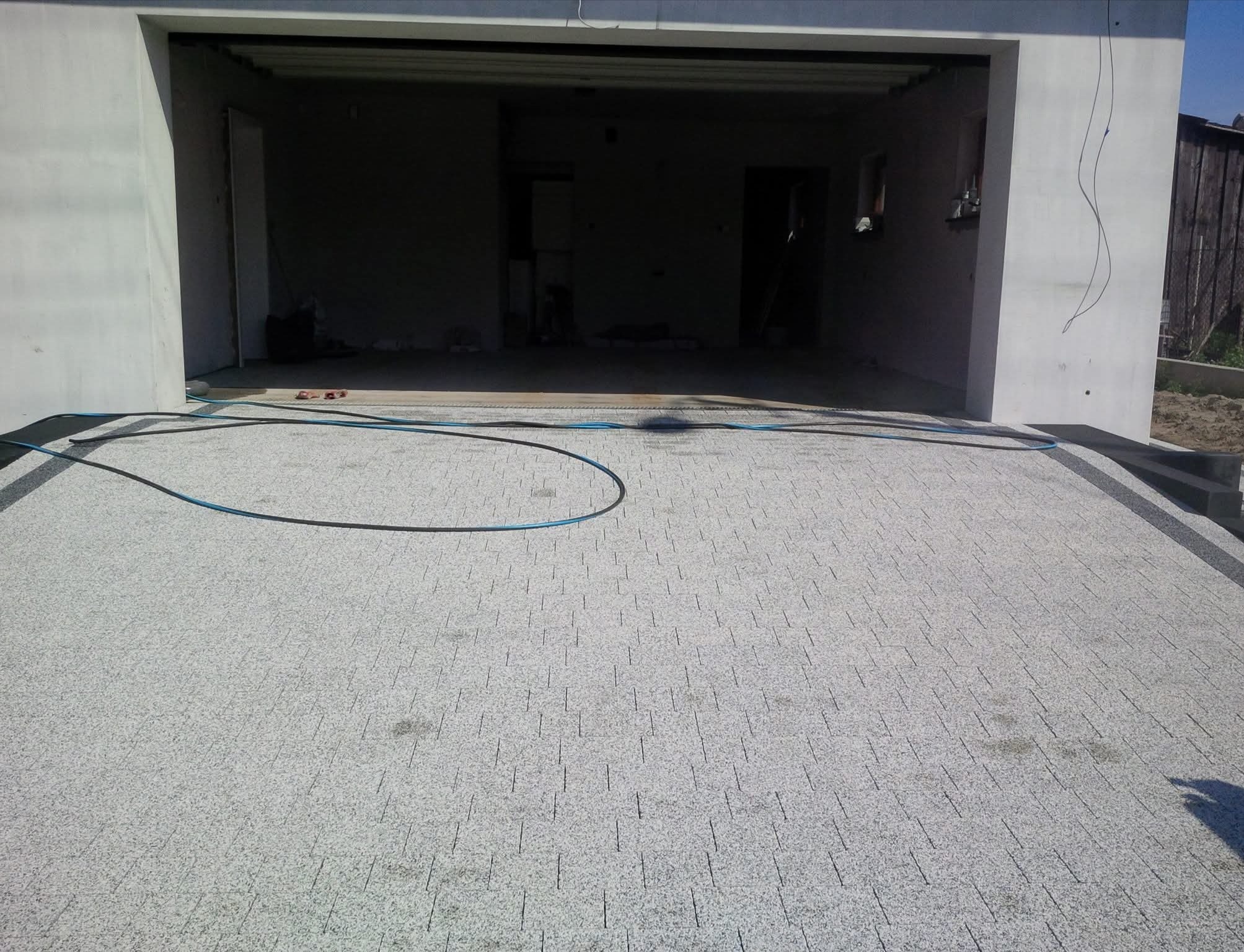 Jasnoszara kostka brukowa przed garażem, z widoczną instalacją w tle. Detal: czarny obrzeże kostki i niebieski przewód na powierzchni. Widok z góry.