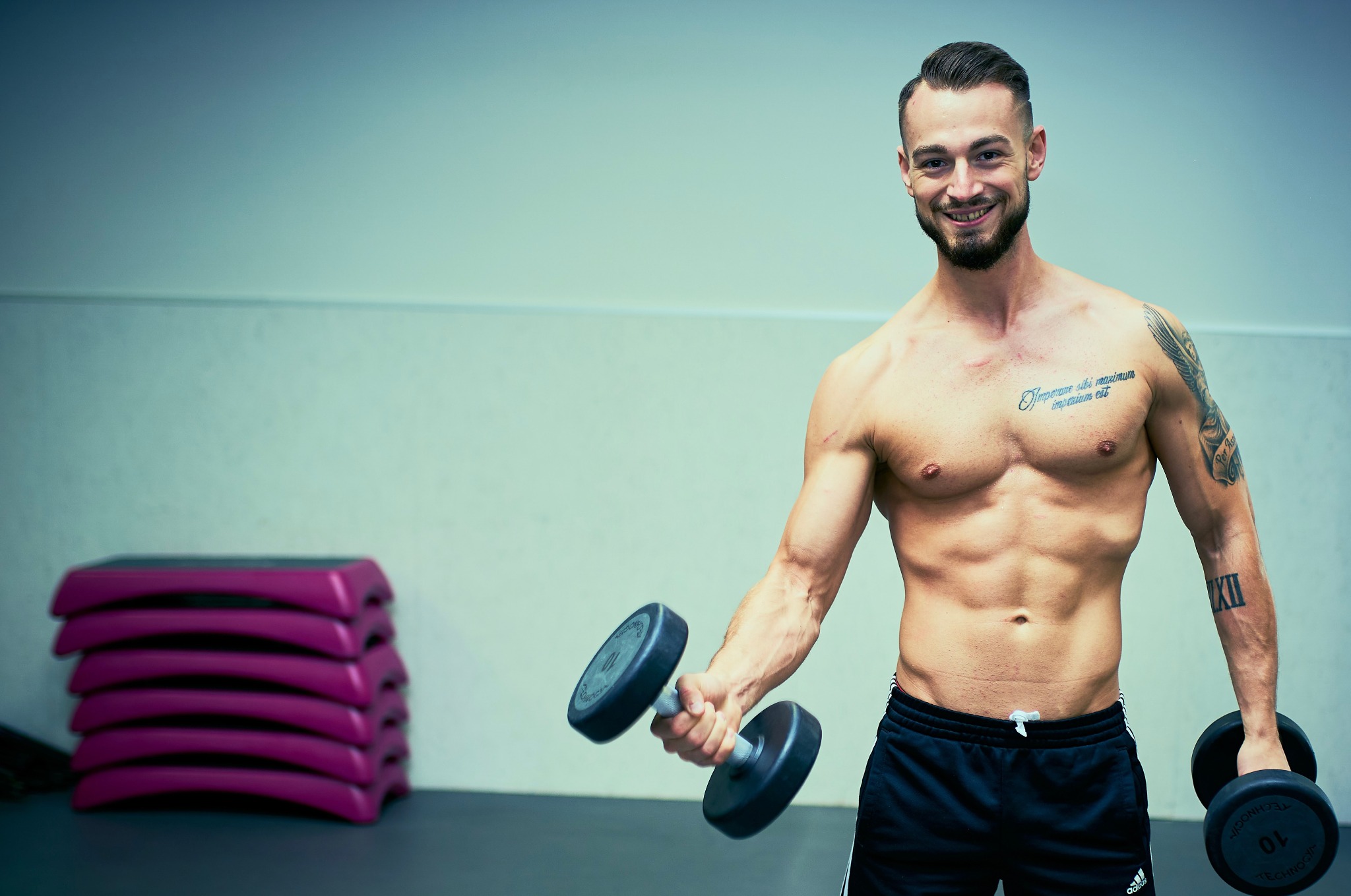 Uśmiechnięty, wysportowany mężczyzna z tatuażami, ćwiczący z hantlami w siłowni. W tle różowe steppery.