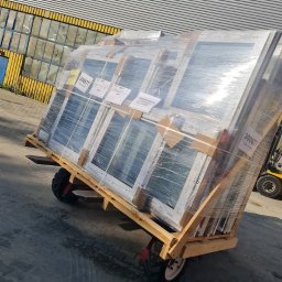 MAK Consulting p.h.u. Michał Nowak - Paleta z oknami PCV, owinięta folią, przewożona na wózku transportowym na zewnątrz budynku o metalowej fasadzie. Widoczne etykiety na oknach.