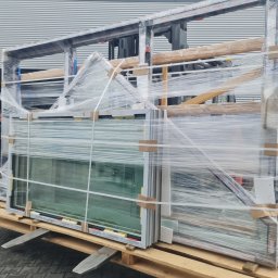 MAK Consulting p.h.u. Michał Nowak - Paleta z zapakowanymi oknami, zabezpieczonymi folią, transportowana wózkiem widłowym na zewnątrz budynku z szarej blachy trapezowej.