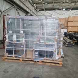 MAK Consulting p.h.u. Michał Nowak - Paleta z zapakowanymi oknami PCV w magazynie, owinięta folią stretch, widoczne kartonowe zabezpieczenia narożników i etykiety producenta.