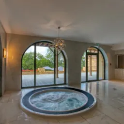 Ekskluzywne wnętrze z jacuzzi i przeszklonymi, łukowymi wyjściami na taras. Marmurowe płytki, dekoracyjne oświetlenie i widok na zieleń.