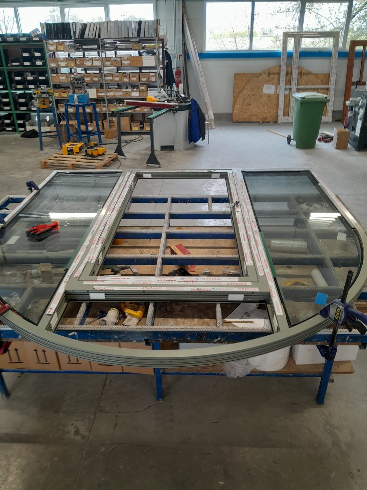 Szkielet okna z łukowym elementem, w warsztacie z narzędziami i materiałami. Widać elementy mocujące i częściowo oszkloną konstrukcję.