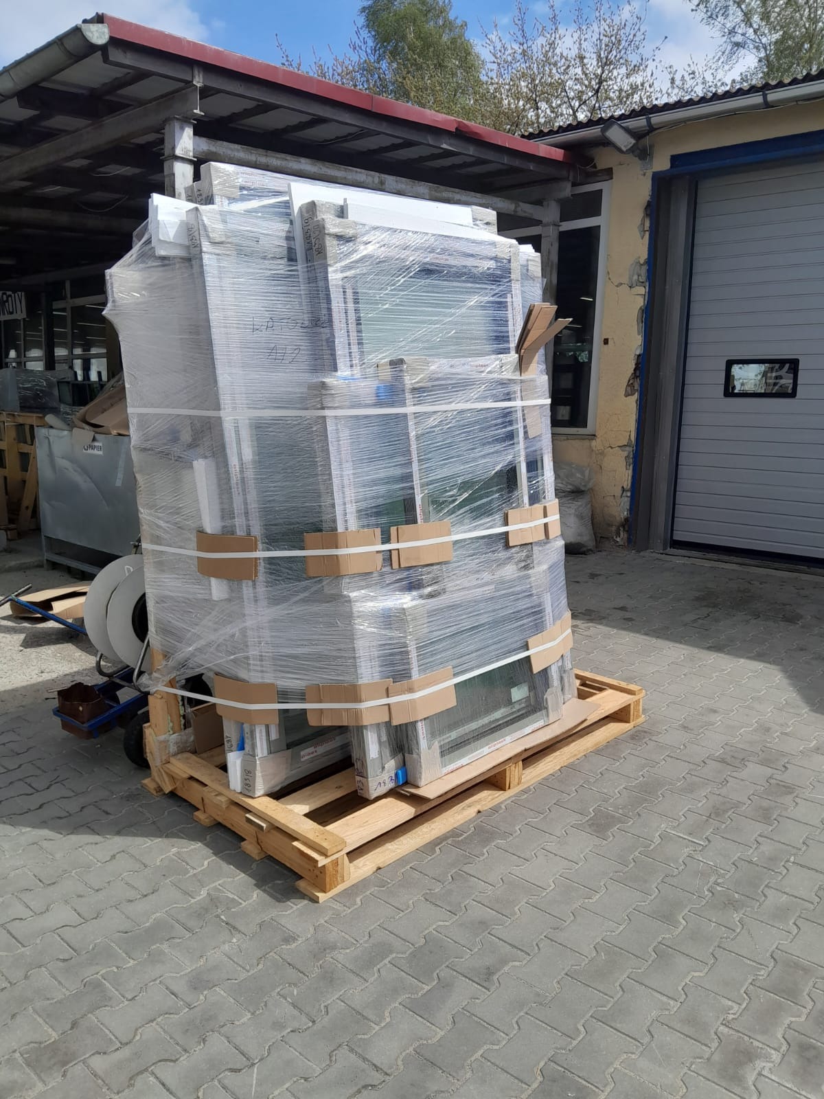 Paleta z oknami zabezpieczonymi folią, przygotowana do transportu. Widoczne kartonowe zabezpieczenia i napisy na oknach. Tło stanowi fragment budynku i kostka brukowa.