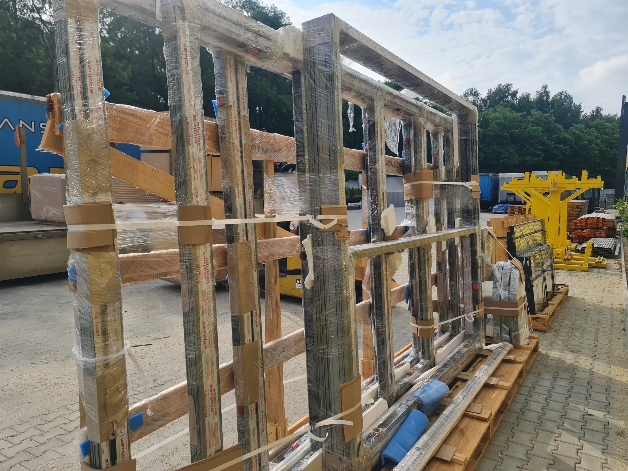 Szkielety okienne Aluplast, zabezpieczone folią i taśmami, ułożone na paletach. Widoczne elementy montażowe i transportowe. Tło: drzewa i błękitne niebo.