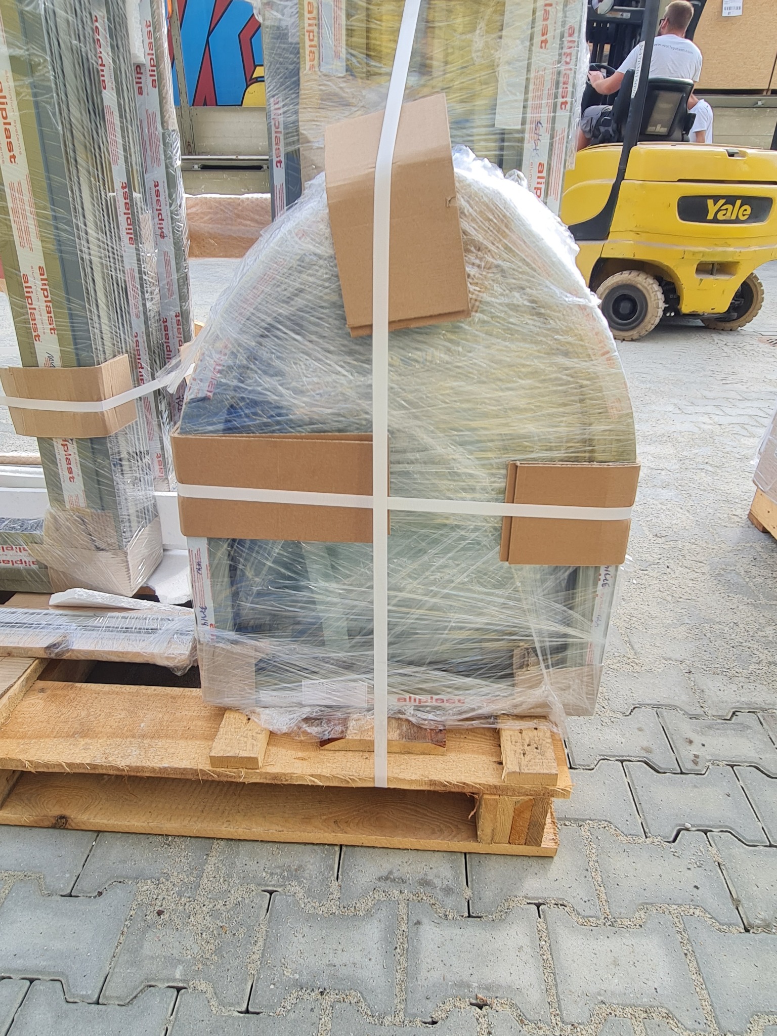 Przygotowane do transportu okno w folii na palecie, z widocznym w tle wózkiem widłowym Yale i innymi oknami Aliplast, oczekujące na montaż.