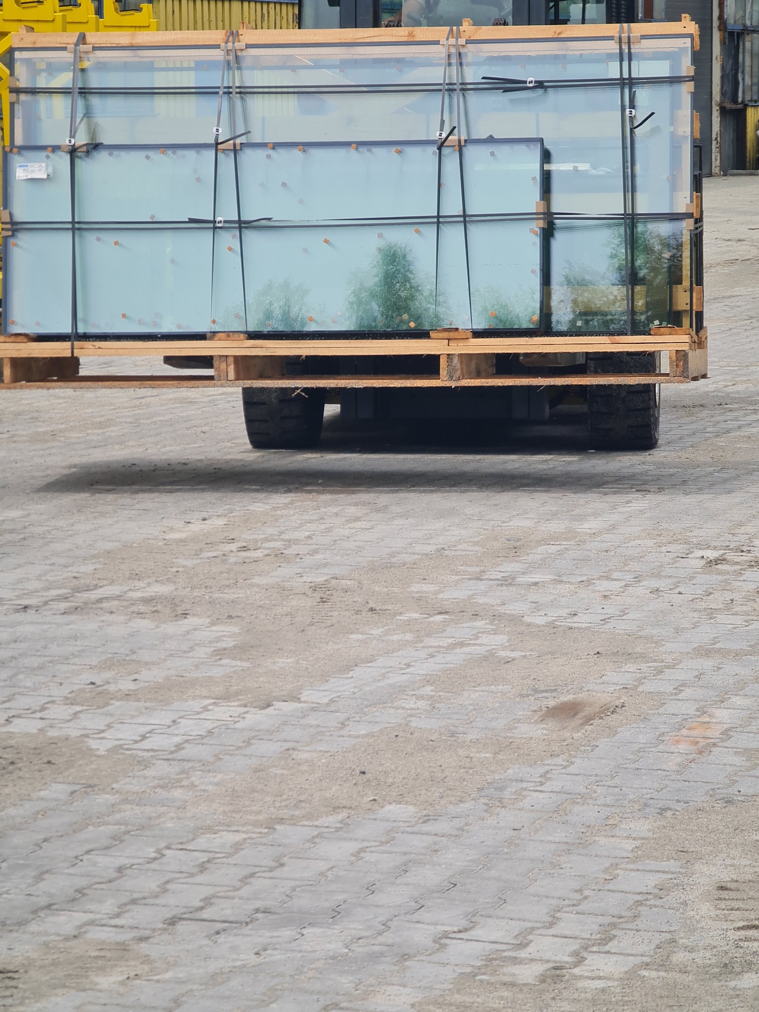 Szyby okienne na palecie transportowane wózkiem widłowym, widoczne zabezpieczenia i refleksy otoczenia na szklanych taflach. Tło stanowi plac z kostki brukowej.