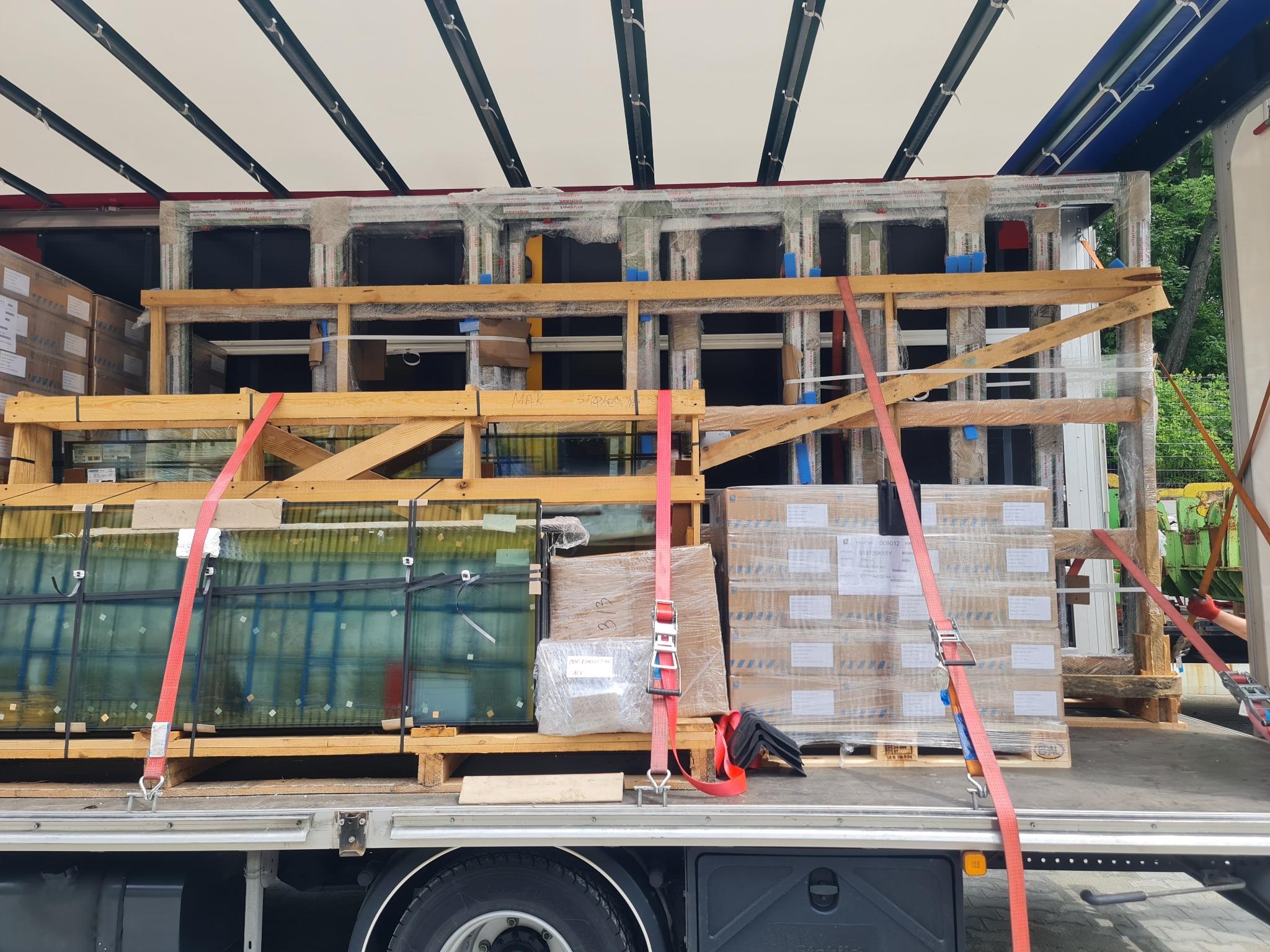 Transport okien i szyb zabezpieczonych pasami na paletach w naczepie ciężarówki. Widoczne skrzynie z oknami, palety i elementy mocujące ładunek.